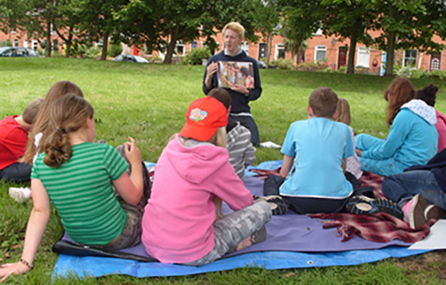 Eine Kindergruppe welche auf einer Wiese auf Decken sitzt und von einem Mann eine Geschichte erzählt bekommt.
