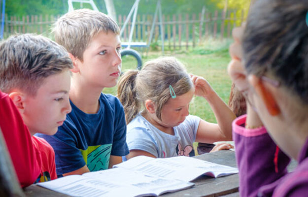 Kinder die an einem Tisch sitzen und offene Heft vor sich liegen haben.