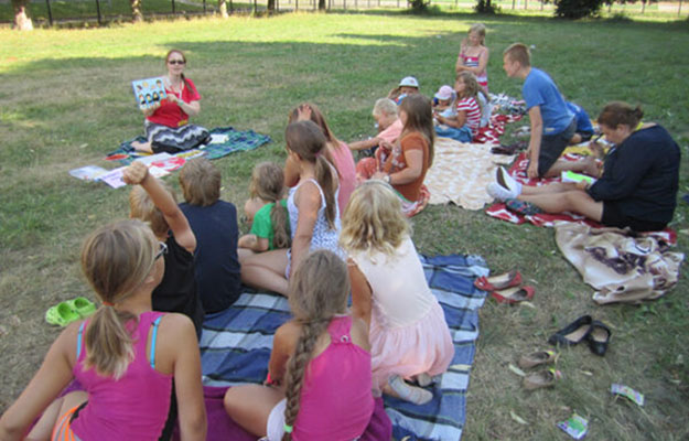 Eine Kindergruppe welche auf einer Wiese auf Decken sitzt und von einer Frau eine Geschichte erzählt bekommt.